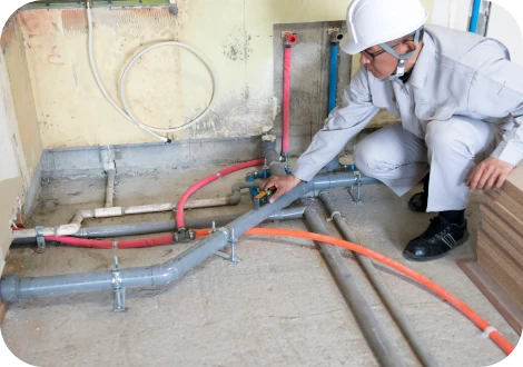 作業服を着た作業員が屋内で配管工事を行っている様子。周囲には工具や配管部品が整然と並び、明るく清潔な作業環境が広がっている。真剣な表情で丁寧に作業している雰囲気が伝わる。