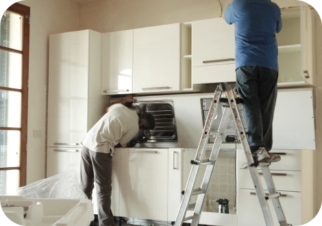 作業服を着た作業員がキッチンのシンク周辺で住宅設備機器の設置作業をしている様子。周囲には工具や部品が整然と並び、明るく清潔な室内環境が広がっている。作業員は丁寧に作業しており、安心感と信頼感が感じられる。