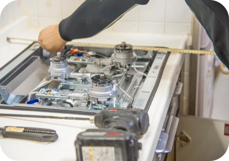 家のガスコンロを整備している作業員の写真。寸法を測り、丁寧な仕事ぶりが伝わる。周囲には工具や部品が整然と並んでいる。